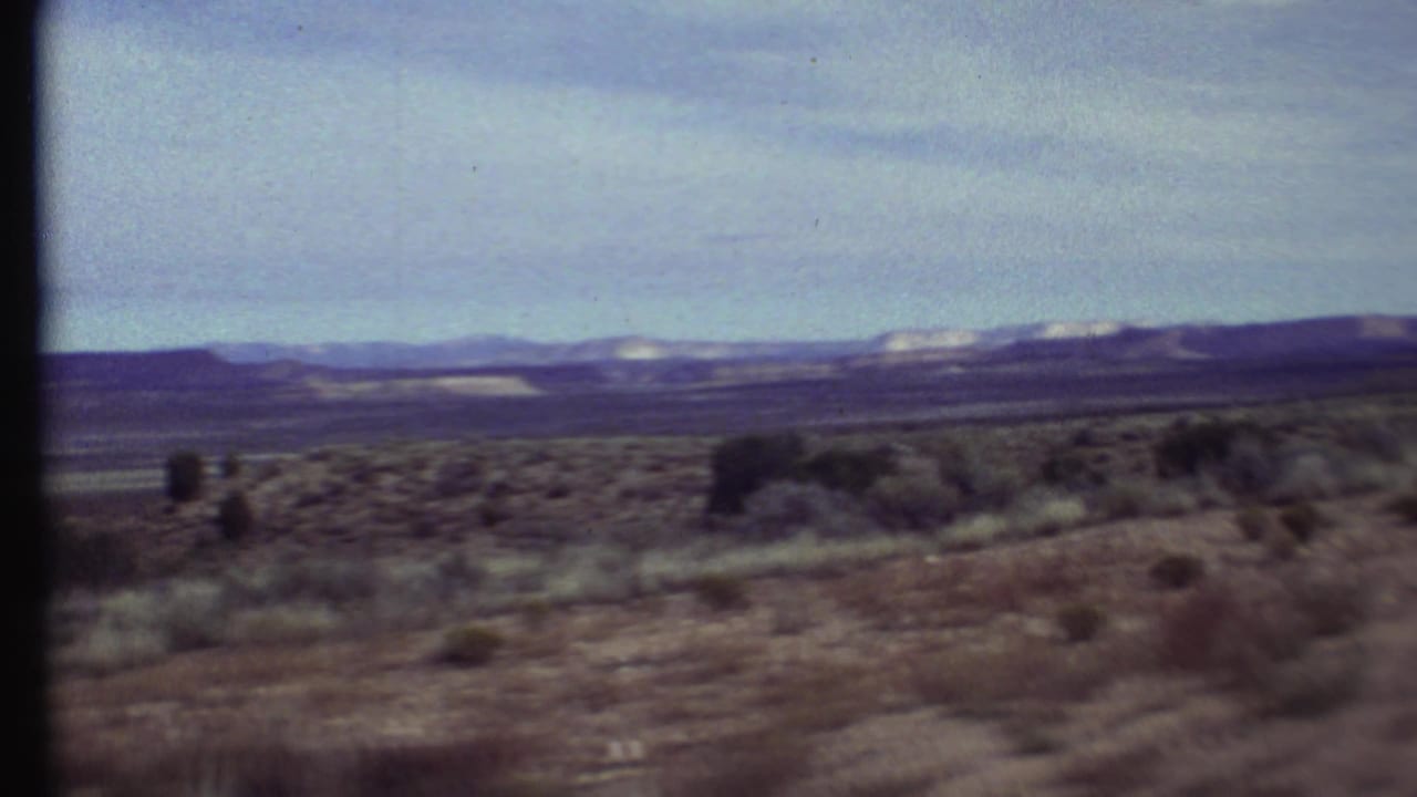 Grand Canyon-region home-movie frame showing scattered desert shrubs, a wide plain, and distant ridgelines beneath a hazy blue sky.