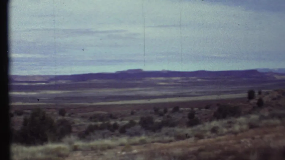 1972 8mm travel view across scrubby Grand Canyon-region terrain toward a long mesa-like horizon under thin clouds.