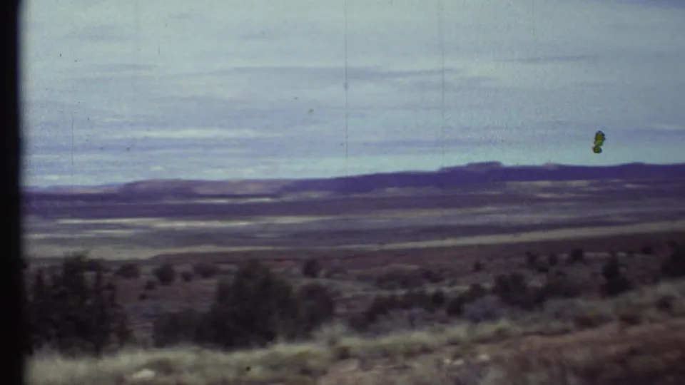 1972 8mm home-movie frame of an arid Arizona overlook with scrubby foreground, distant ridges, and a pale clouded sky.