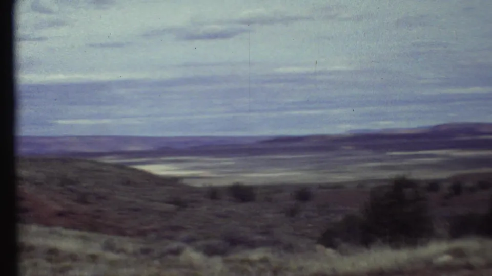 1972 8mm frame from a moving pan across dry Arizona hillside toward a pale valley floor and distant ridges.