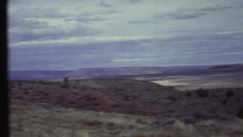 1972 Arizona home-movie frame showing scrubby desert slope, pale valley floor, distant low ridge, and soft cloud cover.