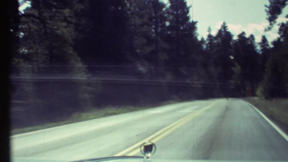 1972 8mm windshield view of a two-lane forest road near Grand Canyon, Arizona, with dashboard and mirror visible.