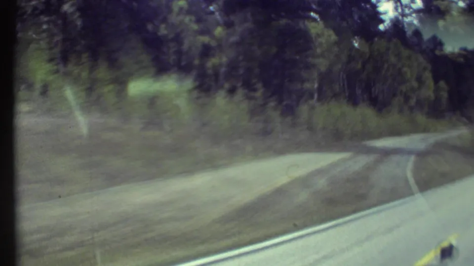 Motion-blurred 1972 8mm windshield view of a wooded Grand Canyon-area road bending past trees and roadside grass.