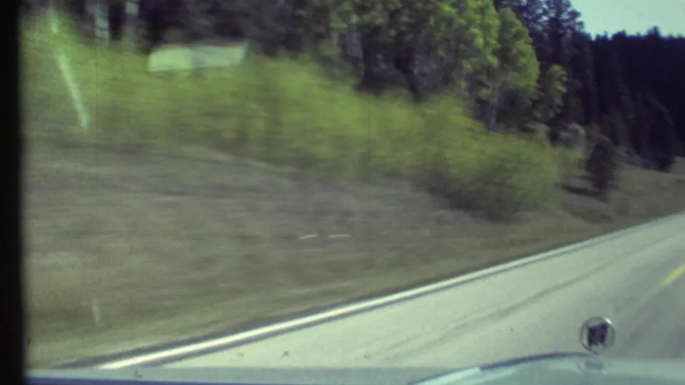 1972 8mm car-window view near Grand Canyon, Arizona, showing a paved road edge, grassy shoulder, and surrounding forest.