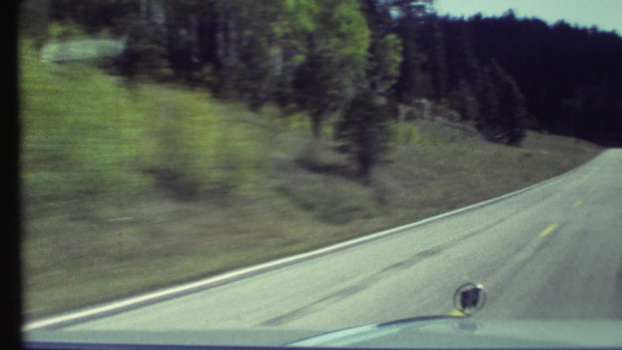 Forward view from a car on a two-lane road curving through grassy hills and trees.