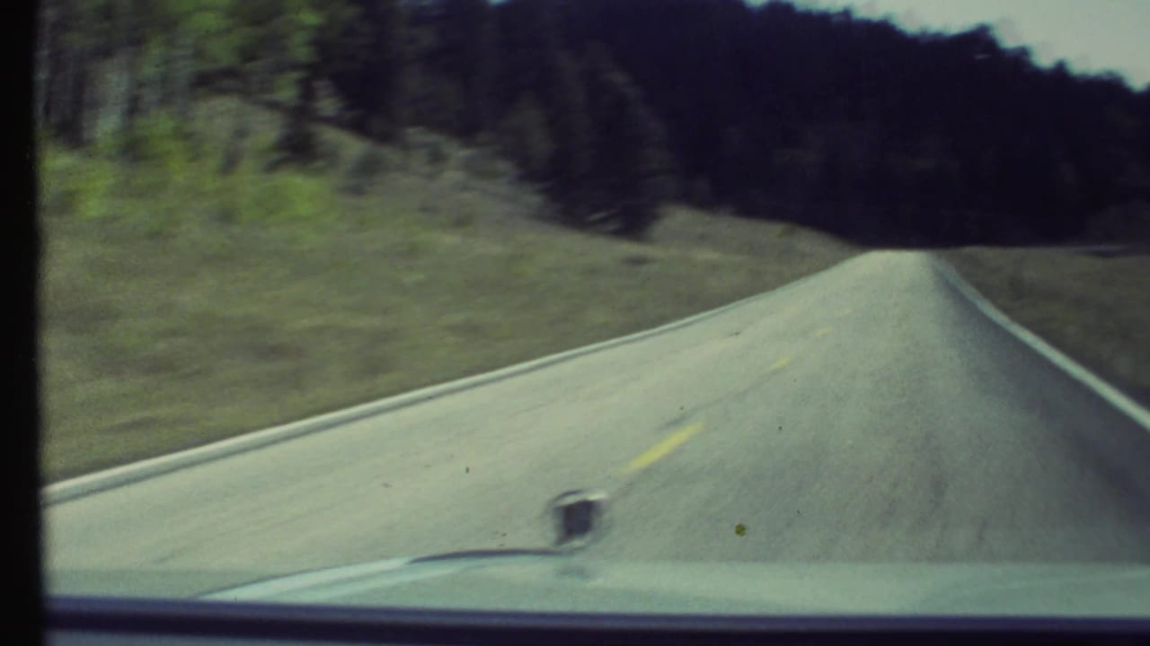 Forward-facing 1972 8mm road view near Grand Canyon, Arizona, with a paved two-lane road, forested hillside, and car hood in frame.