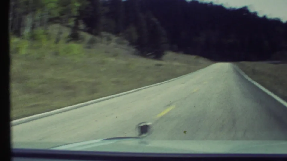 Forward-facing 1972 8mm road view near Grand Canyon, Arizona, with a paved two-lane road, forested hillside, and car hood in frame.