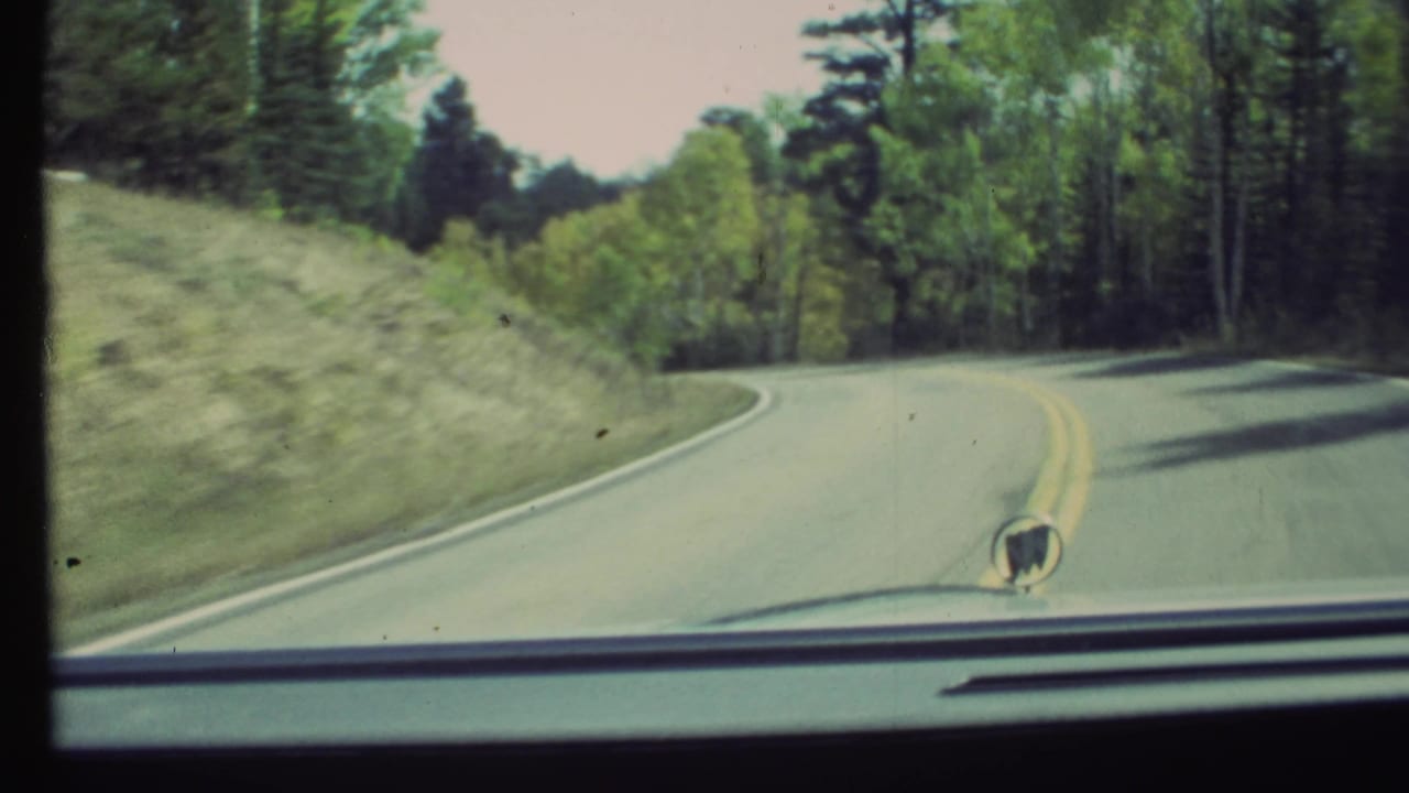 1972 8mm car POV near Grand Canyon, Arizona, with a curving road, yellow lines, forested roadside, dashboard edge, and hood ornament.