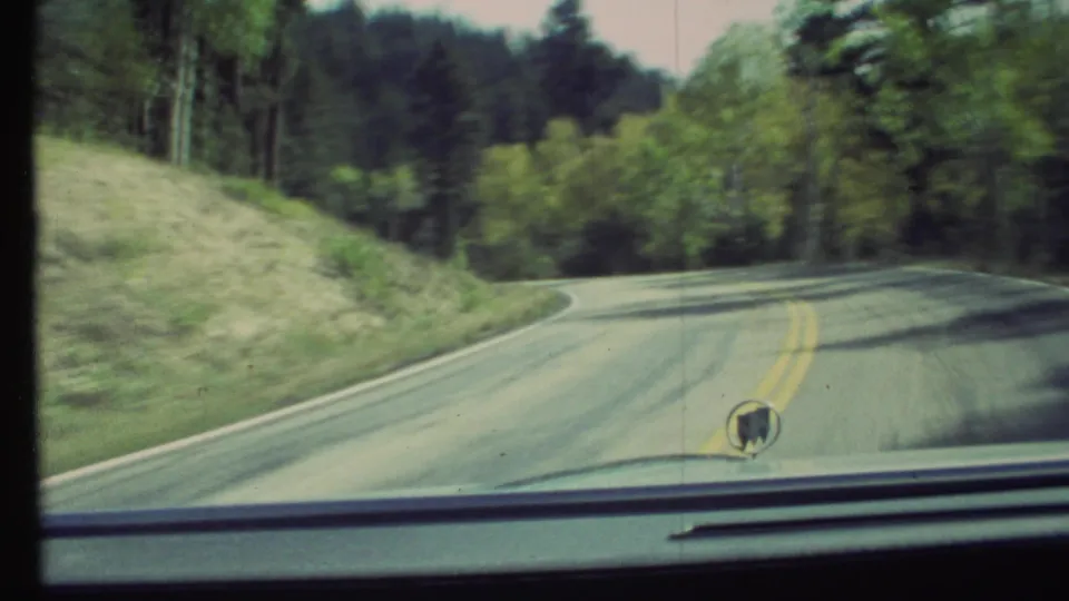 Vintage 8mm car POV from 1972 showing a shaded curve in a two-lane road with yellow lines, grassy shoulder, trees, and hood ornament.