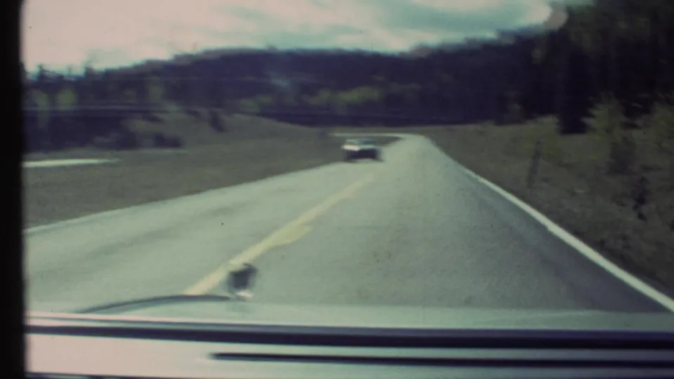 Soft 8mm frame from a moving car on a Grand Canyon, Arizona area road, with a distant vehicle, open hills, roadside trees, and visible dashboard edge.