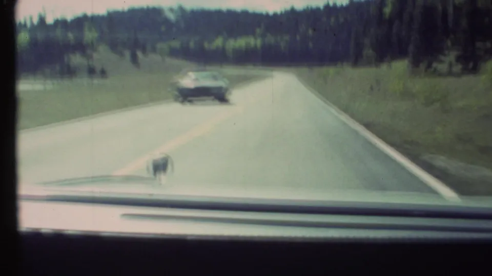 1972 8mm car POV on a rural approach road near Grand Canyon, with a sedan passing and forested hills ahead.