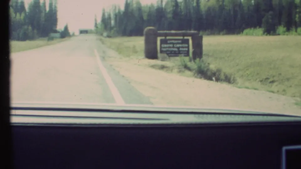 1972 8mm windshield view approaching the Grand Canyon National Park entrance sign on a two-lane road in Arizona.