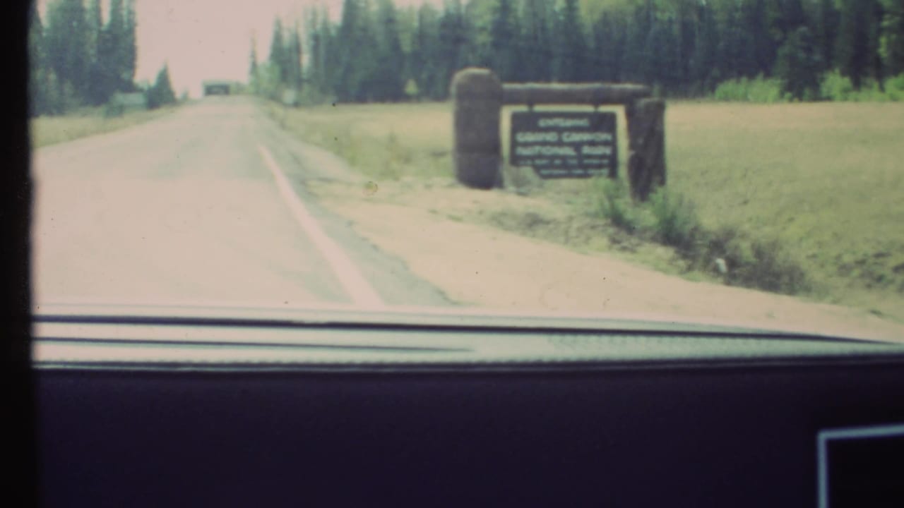 Windshield view of the Grand Canyon National Park entrance sign beside a forested road, with soft 1972 8mm color and blur.