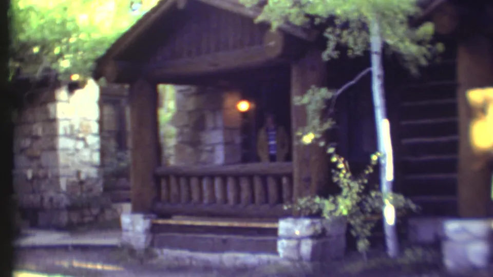 Rustic Grand Canyon-area cabin or lodge porch in 1972 footage, with stone foundation, wood railing, trees, and a person silhouetted in the doorway.