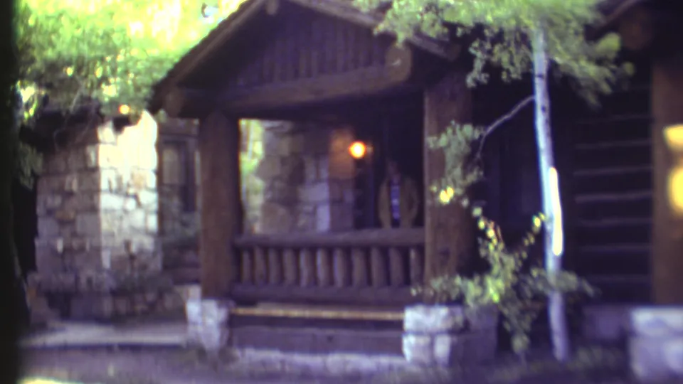 Soft 8mm frame of a rustic cabin porch near the Grand Canyon, showing a doorway figure, wood logs, stonework, foliage, and warm interior light.