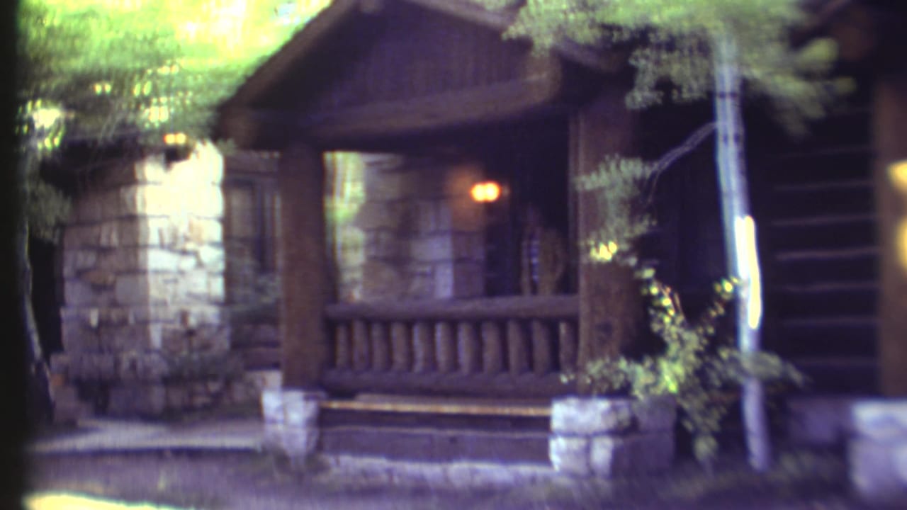 Rustic log-and-stone building with porch railing, trees, and a glowing exterior light.
