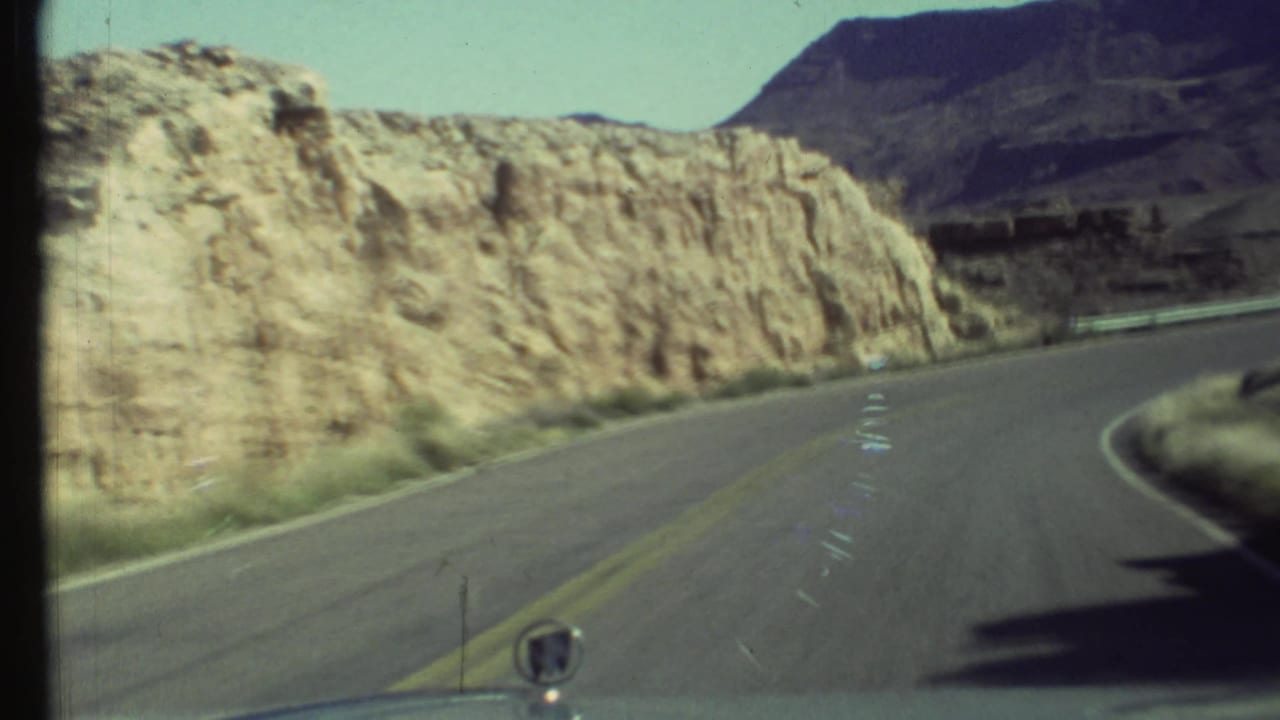 1972 Grand Canyon road view from inside a moving vehicle, with a curving highway, pale rock cut, and dark mesa in the distance.