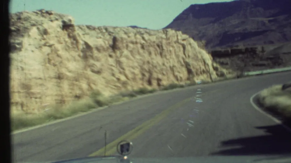 1972 Grand Canyon road view from inside a moving vehicle, with a curving highway, pale rock cut, and dark mesa in the distance.