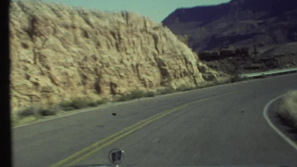 Vehicle-mounted 8mm view of a Grand Canyon-area highway bending alongside tall pale rock formations under a clear sky.