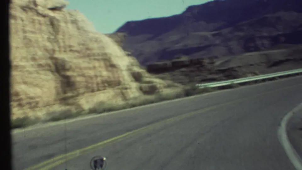 Soft 1972 8mm frame from a moving vehicle, showing a Grand Canyon road curve, guardrail, pale rock face, and darker canyon slopes.
