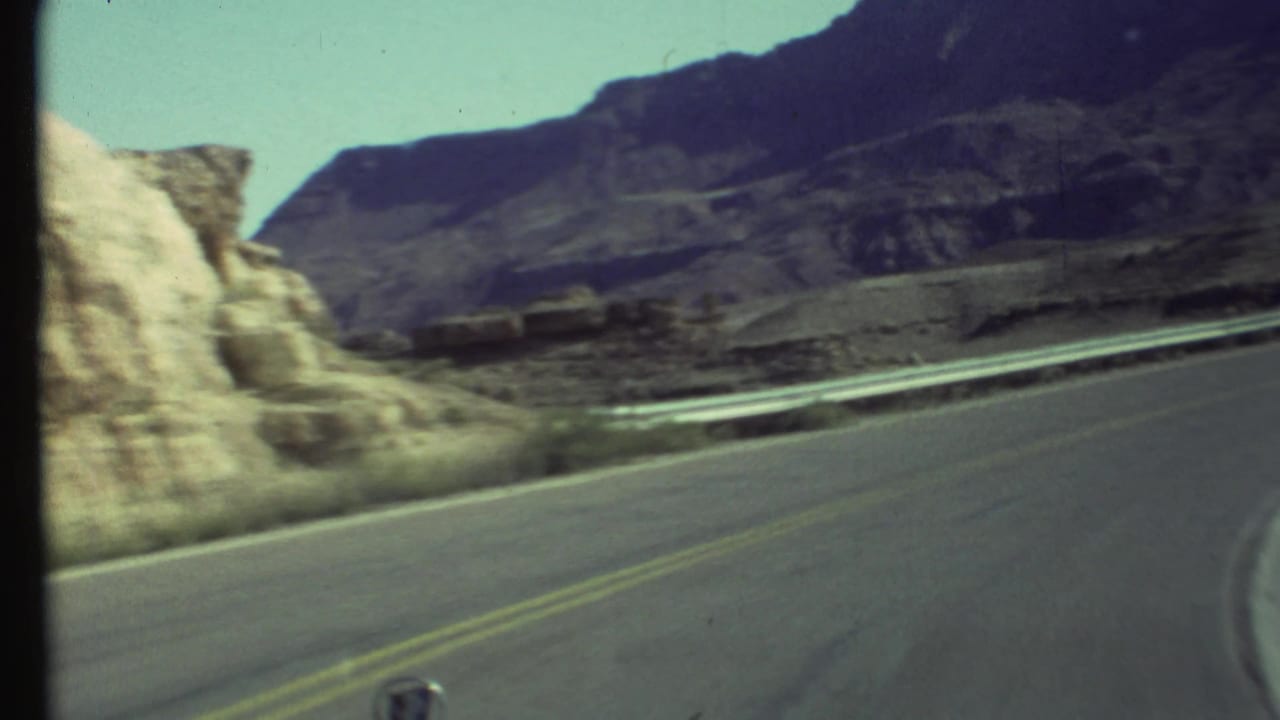 Curving paved mountain road with guardrail, rocky slope, and dark canyon ridges beyond.