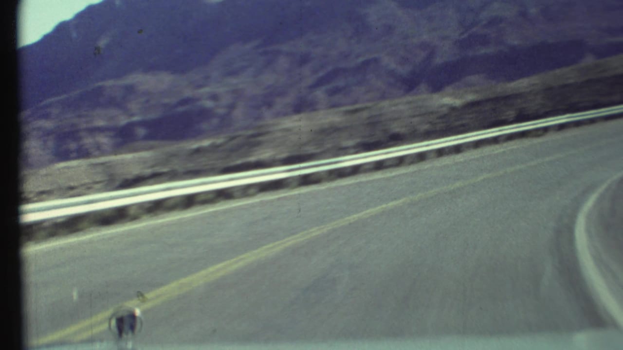 Paved road bending right beside a guardrail with rugged mountains in the distance.
