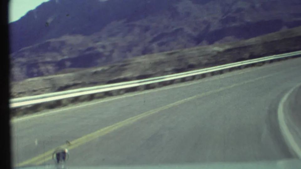 Passenger-side 8mm view of a right-curving Grand Canyon area road, metal guardrail, and rugged desert mountains under clear sky.