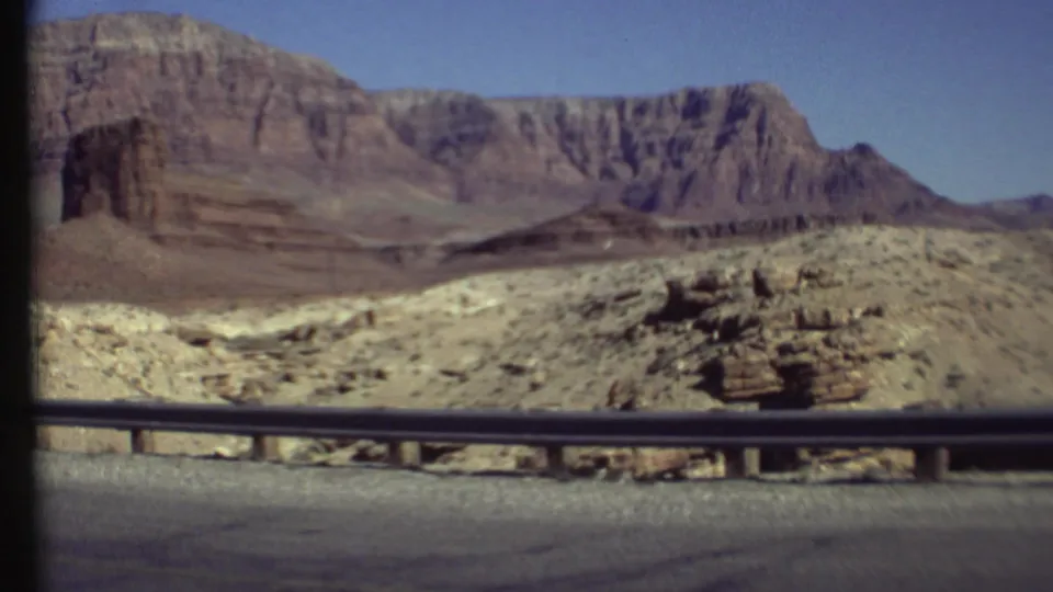 1972 Grand Canyon home-movie frame showing layered cliffs, pale rocky ground, sparse vegetation, and a roadside guardrail from a moving car.