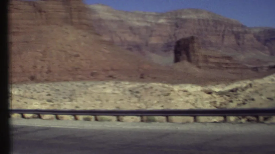 1972 8mm view of layered red rock cliffs in the Grand Canyon area, seen from a moving roadside perspective.