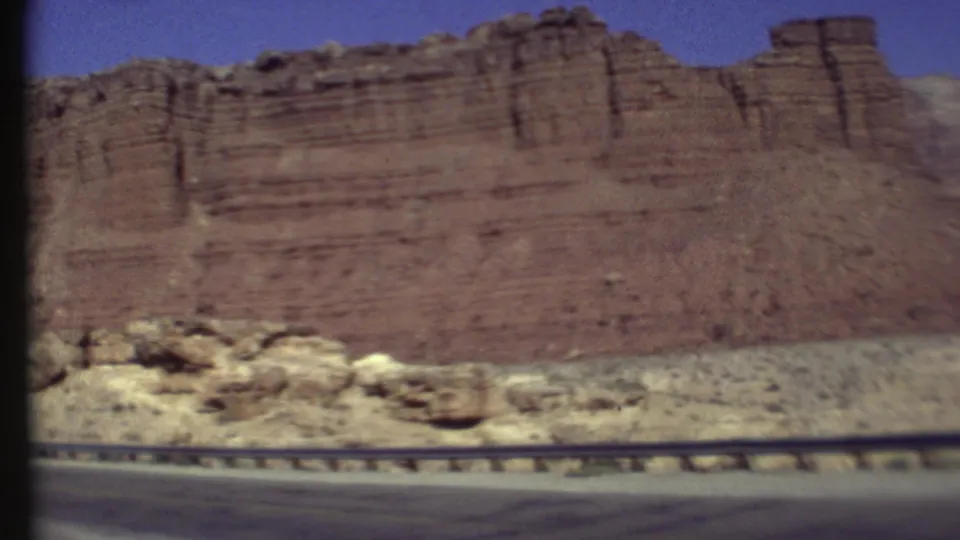 1970s 8mm frame showing layered Arizona red rock cliffs, roadside rocks, and a highway guardrail from a moving vehicle.