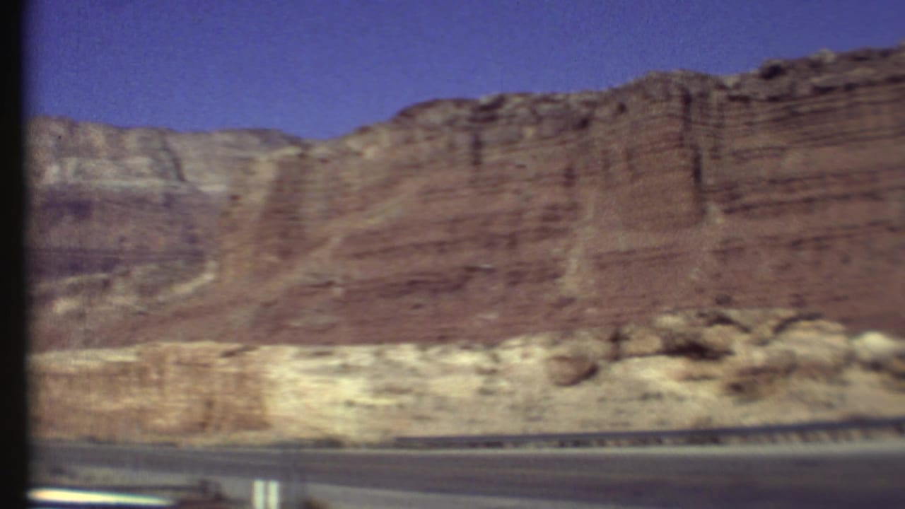 1972 8mm Grand Canyon road-trip frame showing reddish layered cliffs and pale rock bands beneath a clear blue sky.