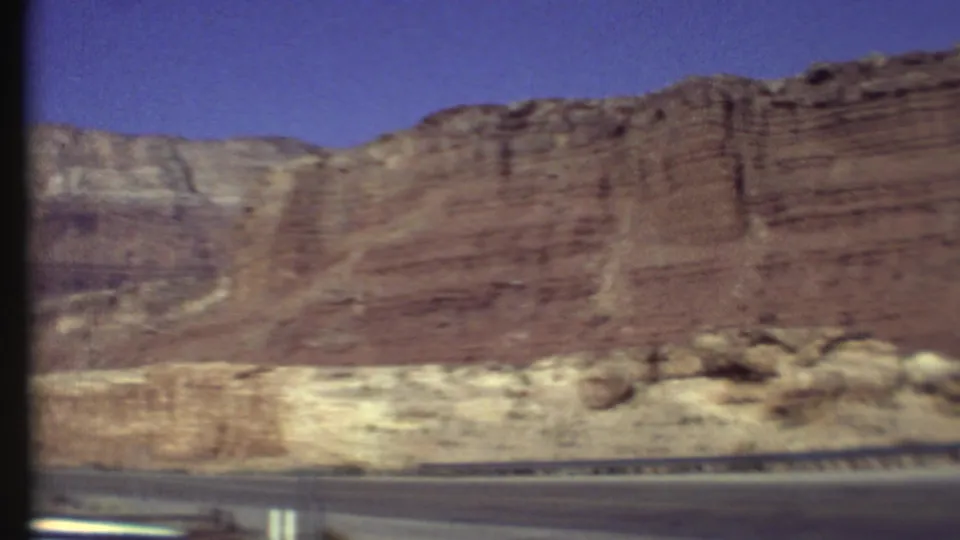 1972 8mm Grand Canyon road-trip frame showing reddish layered cliffs and pale rock bands beneath a clear blue sky.