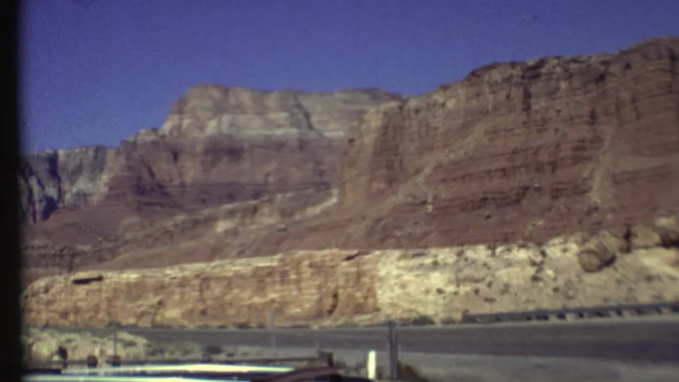 Grand Canyon, Arizona home-movie frame with stratified cliffs, a paved roadside edge, and blue sky from a moving car viewpoint.