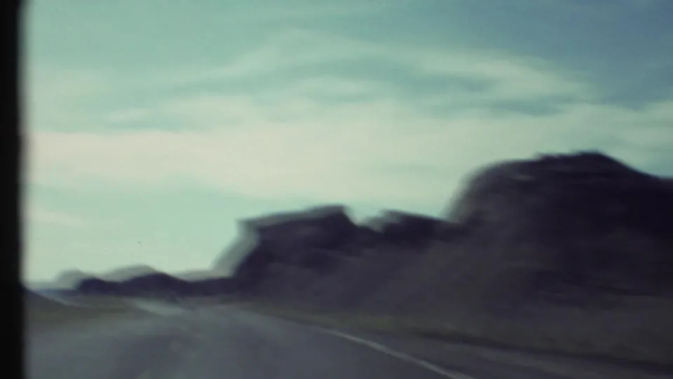 Moving 8mm road-trip frame showing a desert highway curving past dark canyon rock forms, with blur from filming through travel motion.