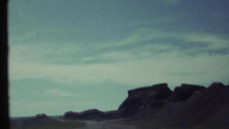 1972 Grand Canyon-area road view with low desert mesas, dark rock silhouettes, and an open sky captured on soft 8mm film.