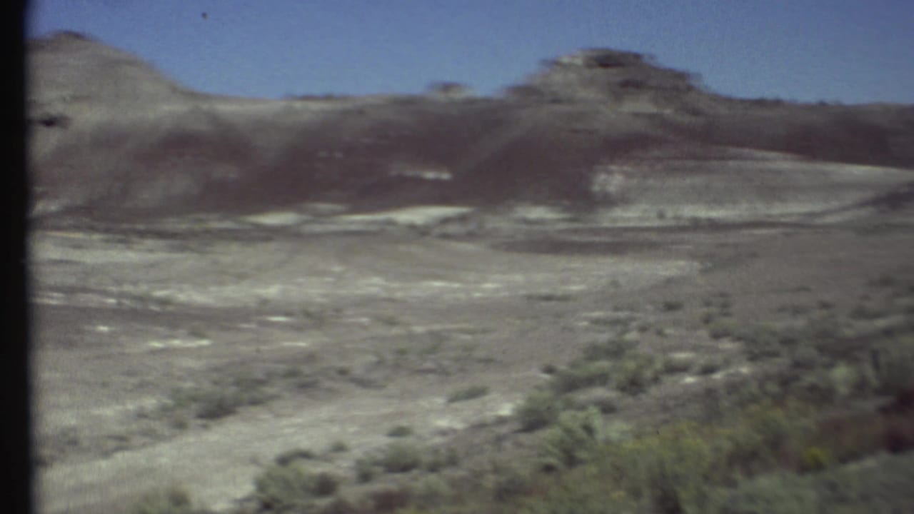 Wide desert basin with sparse shrubs and eroded rocky hills under blue sky.