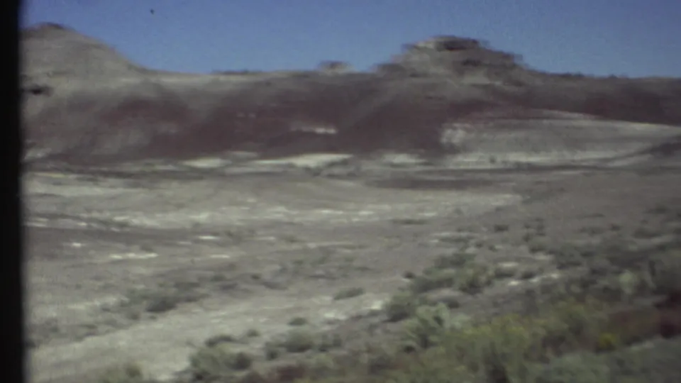 1972 8mm view near Grand Canyon, Arizona, showing a pale desert basin, scattered scrub, and layered rocky hills.