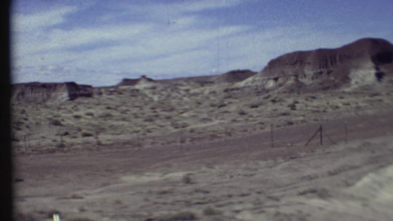 1972 8mm Grand Canyon area view of arid ground, scattered scrub, distant eroded hills, and a blue sky with thin clouds.