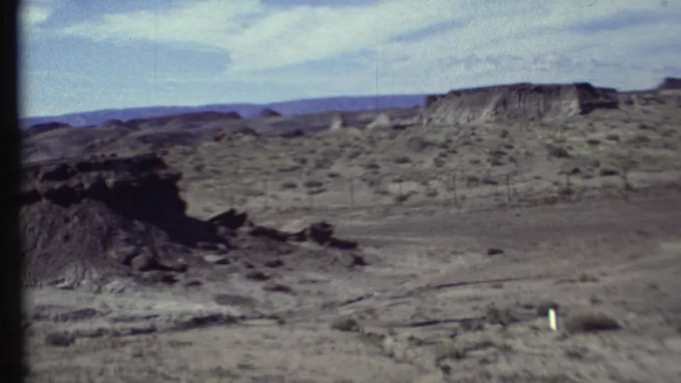 1972 Arizona travel footage frame with an eroded slope in the foreground and scrubland leading toward a distant plateau.