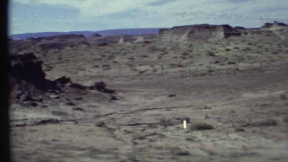 Grand Canyon area 8mm frame showing dry foreground soil, scattered desert shrubs, a low mesa, and soft handheld film texture.