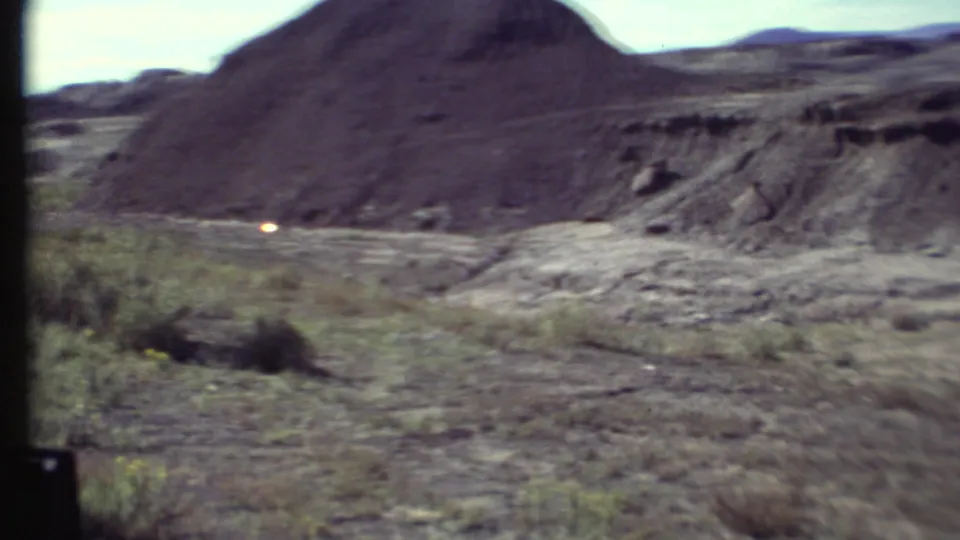 1972 8mm desert view near the Grand Canyon showing a dark slope, pale eroded terrain, and sparse foreground vegetation.