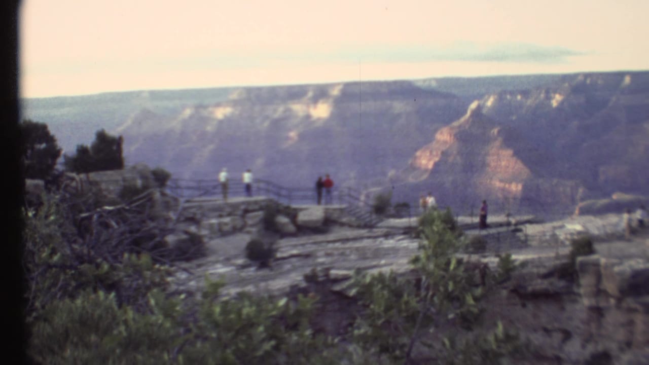 Tourists stand near a canyon overlook railing with layered cliffs in the distance.