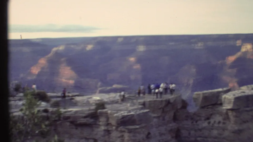 1972 Grand Canyon home-movie frame with tourists on a stone overlook, shrubs in front, and layered cliffs across the gap.