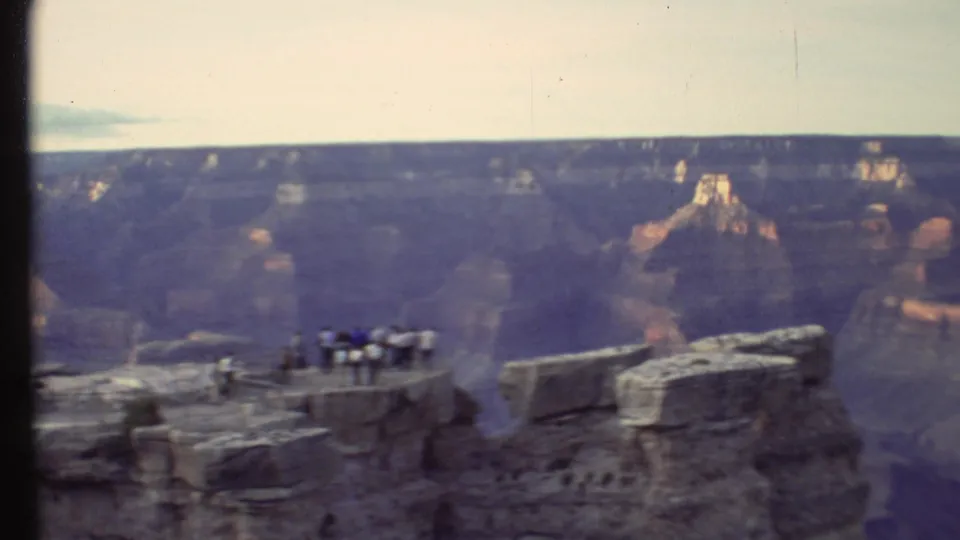Wide 8mm view of tourists at a Grand Canyon overlook in 1972, framed by rocky ledges and distant sunlit formations.
