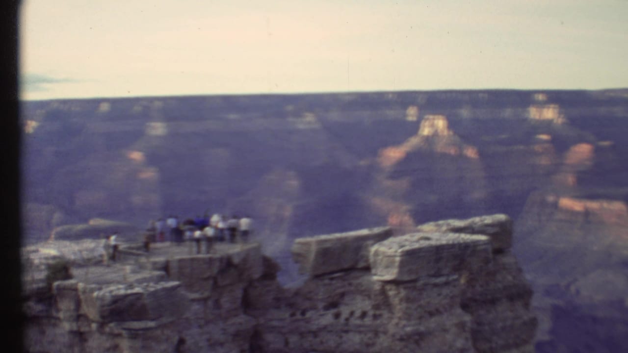 Tourists stand on a rocky Grand Canyon overlook with layered canyon walls beyond.