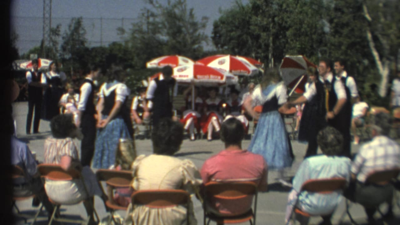 Couples in folk dress dance outdoors before a seated audience under red and white umbrellas.