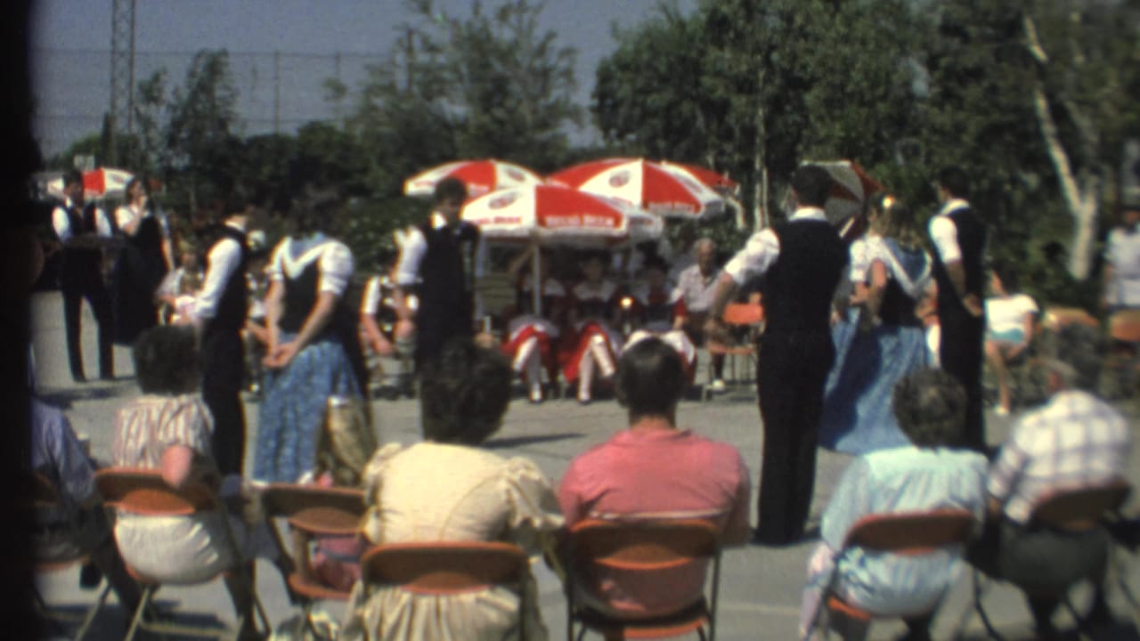 Dancers in vests and blue skirts move in front of spectators and sun umbrellas.