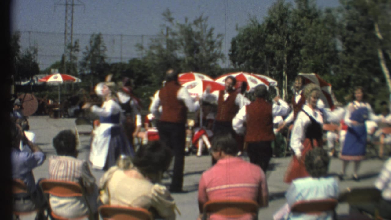 Costumed dancers move across a paved outdoor performance area before spectators.