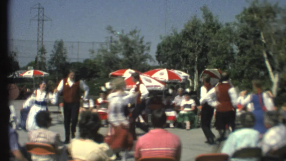 The 1972 8mm footage shows a central male folk dancer with other performers moving past and spectators seated behind them.