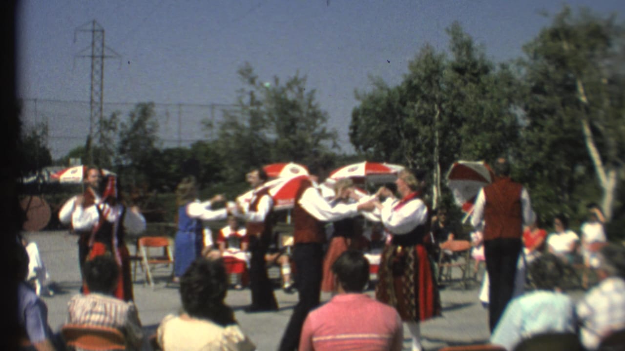 Costumed folk dancers perform outdoors while spectators sit in folding chairs.